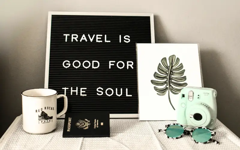 A flat lay composition featuring a travel quote board reading “Travel is good for the soul,” alongside a passport, camera, sunglasses, mug, and framed leaf art — symbolizing wanderlust and self-discovery.